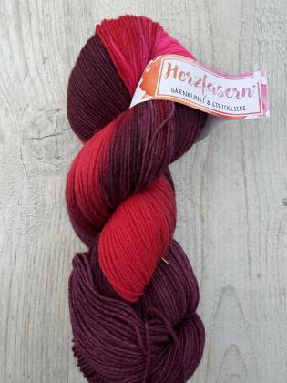 Ein handgefärbter Garnstrang in tiefen Rot- und Beerentönen mit Akzenten in Pink liegt auf einem hellen Holzuntergrund. Die Farben verlaufen weich ineinander und wirken kräftig und zugleich harmonisch.